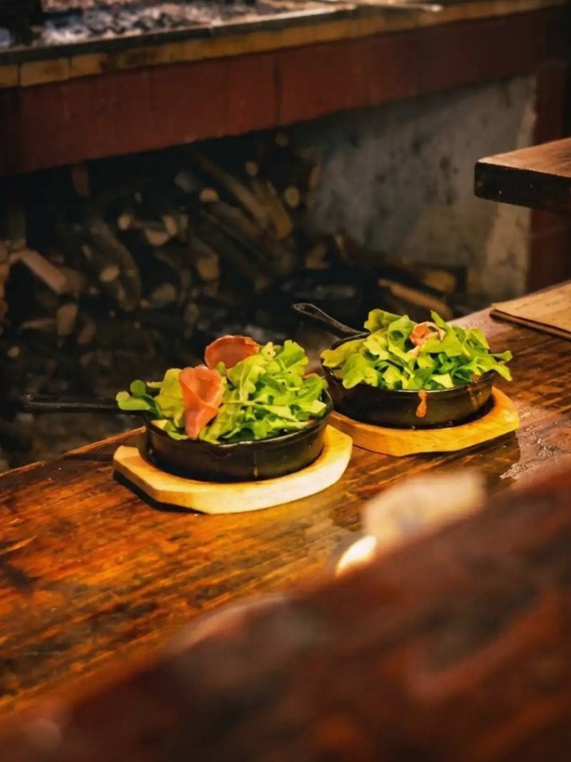 Dos ensaladas en cazuelas de hierro sobre tablas de madera, con la leña de la parrilla detrás